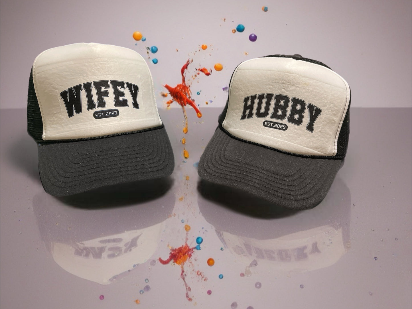 Two trucker hats, one labeled 'Wifey' and the other 'Hubby', on a reflective surface with colorful splashes.