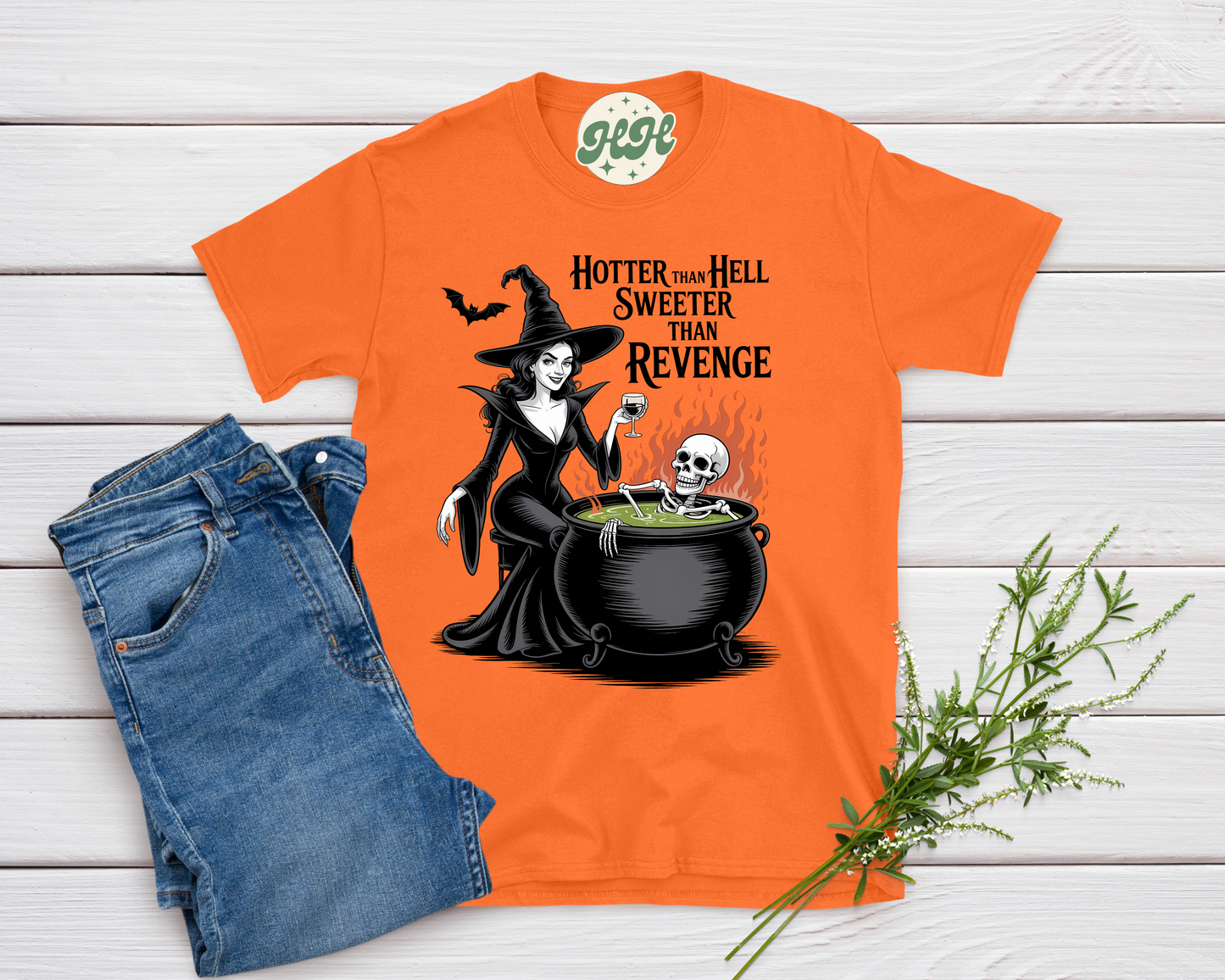 Orange t-shirt with Halloween-themed design featuring a witch and text, paired with blue jeans on a wooden surface.