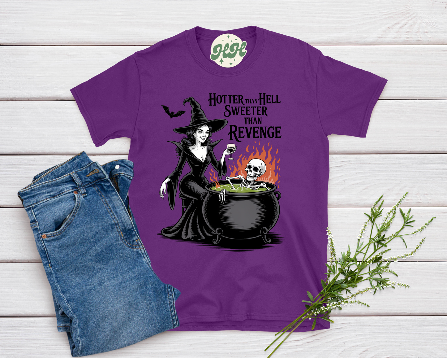Purple t-shirt with witch graphic and text on a wooden surface with jeans and a plant.