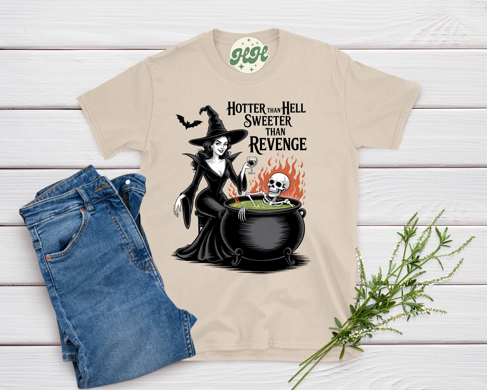 Beige t-shirt with a witch graphic and text, paired with blue jeans on a wooden surface.