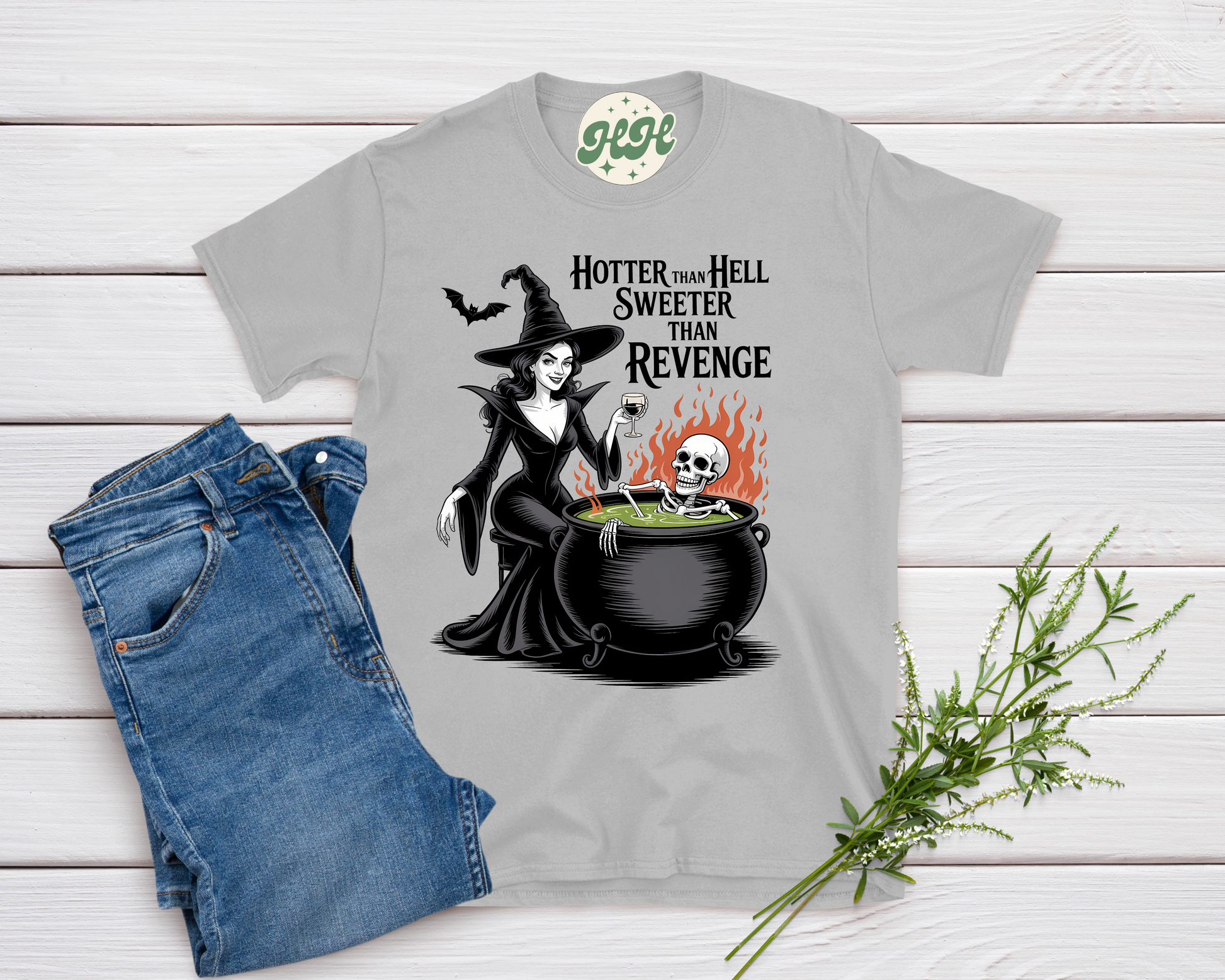 Gray t-shirt with witch graphic and text 'Hotter than Hell Sweeter than Revenge' on a wooden surface with jeans and a plant.