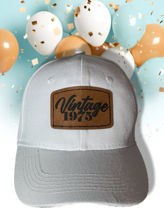White cap with brown 'Vintage 1975' patch on a light blue background with balloons and confetti.