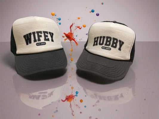 Two trucker hats, one labeled 'Wifey' and the other 'Hubby', on a reflective surface with colorful splashes.