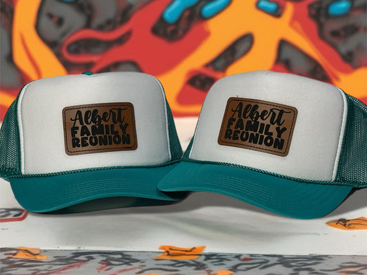 Two white and teal trucker hats with 'Albert Family Reunion' patches on a colorful background.