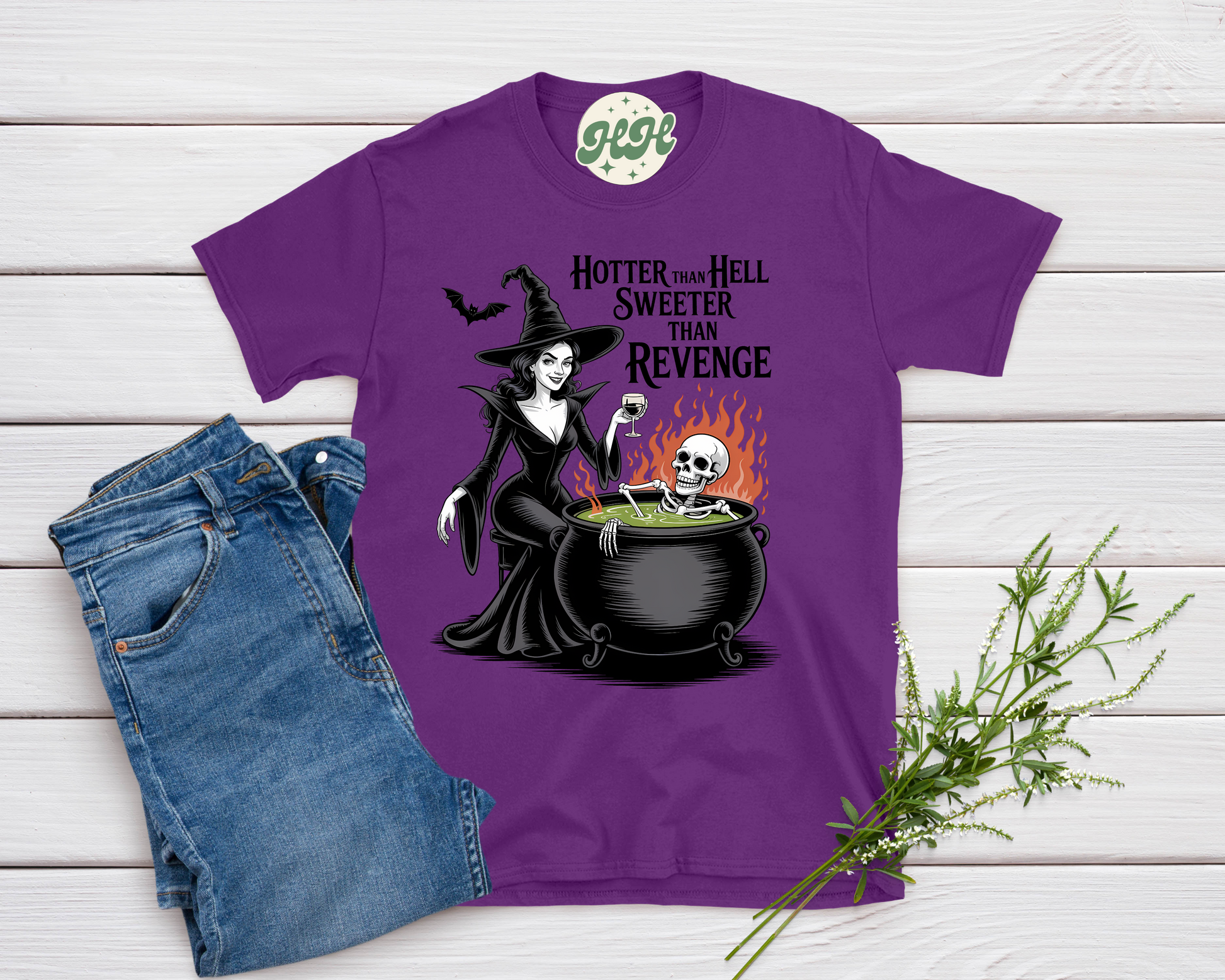 Purple t-shirt with witch graphic and text on a wooden surface with jeans and a plant.