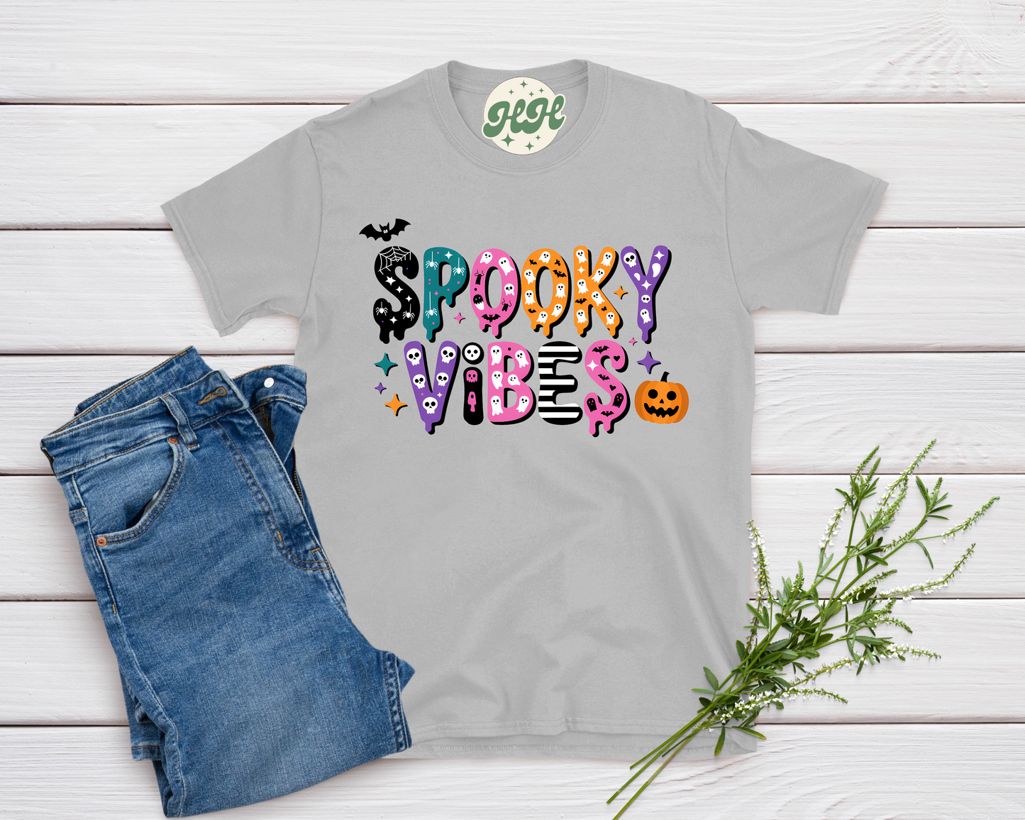 Gray t-shirt with 'Spooky Vibes' design on a wooden surface with jeans and a plant.